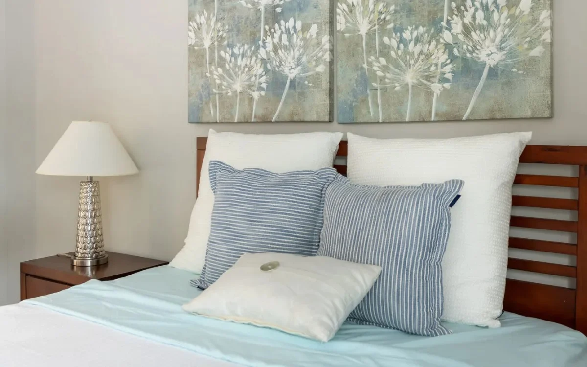 A cozy bedroom scene featuring a neatly made bed with white and striped pillows, a light blue bedspread, and a modern lamp on a bedside table. Above the bed are two pieces of artwork depicting floral patterns in muted tones.