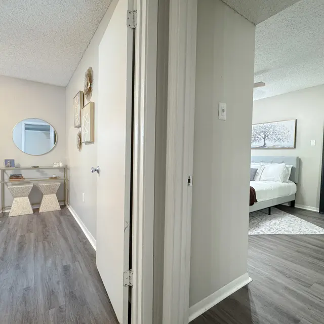 View of a hallway leading to two bedrooms, with light wooden flooring, a mirror on the wall, and one room featuring a bed and decorative artwork.