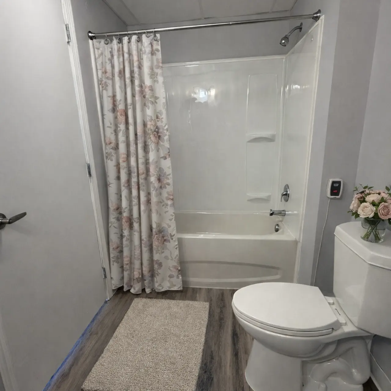 A modern bathroom featuring a white bathtub with a floral shower curtain, a toilet, and a small rug on the wooden floor. A small vase with flowers is placed on the toilet tank.