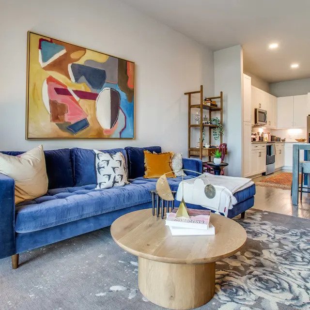A stylish modern living room featuring a blue velvet sofa with various decorative pillows, a round wooden coffee table, and an abstract painting on the wall. The space includes a light-colored rug, a floor lamp, and a kitchen area in the background with white cabinets and appliances.