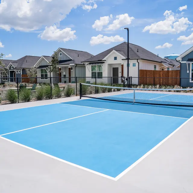 A bright blue tennis court surrounded by residential houses. The court is well maintained with a net in the center, and a clear sky with clouds above.