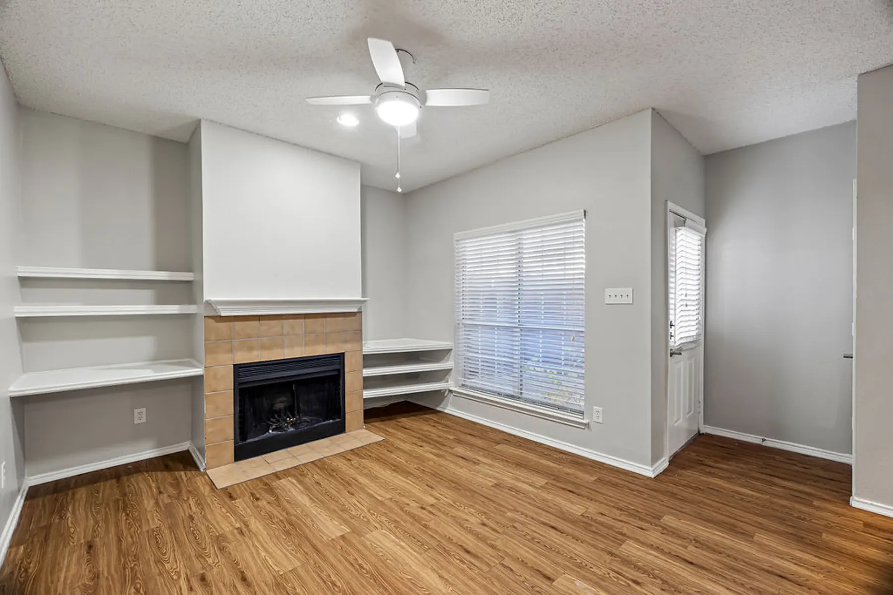 Cozy Living Room Design A cozy living room featuring light-colored walls, hardwood flooring, a ceiling fan, a fireplace with a polished stone facade, and built-in shelves on one side. A window allows natural light to enter the space, and a door leads to another area.