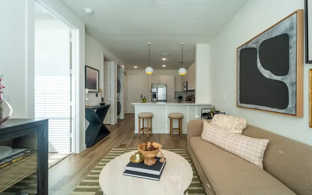 Stylish Modern Living Room A stylish modern living room featuring a beige sofa, a round coffee table, and an open kitchen area. The room is decorated with wall art and vibrant light fixtures.