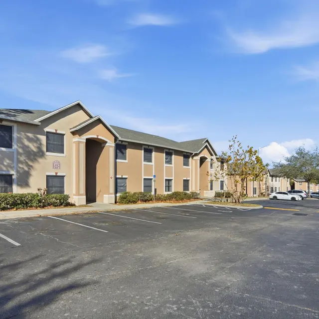 View of a residential apartment building with a parking lot and trees, under a clear blue sky.