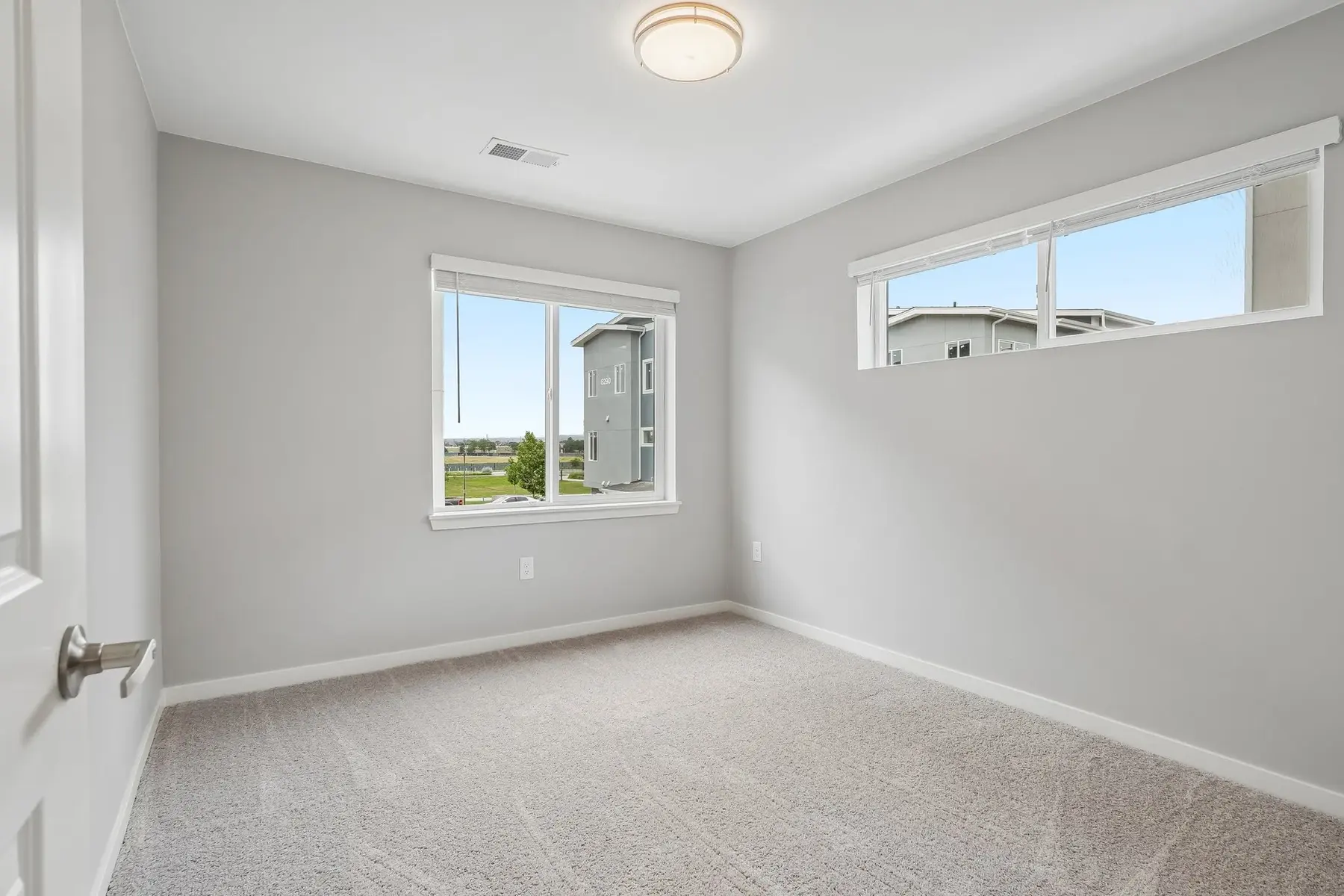 Bright Vacant Bedroom A vacant bedroom with light gray walls and carpet, featuring two windows that allow natural light to enter.