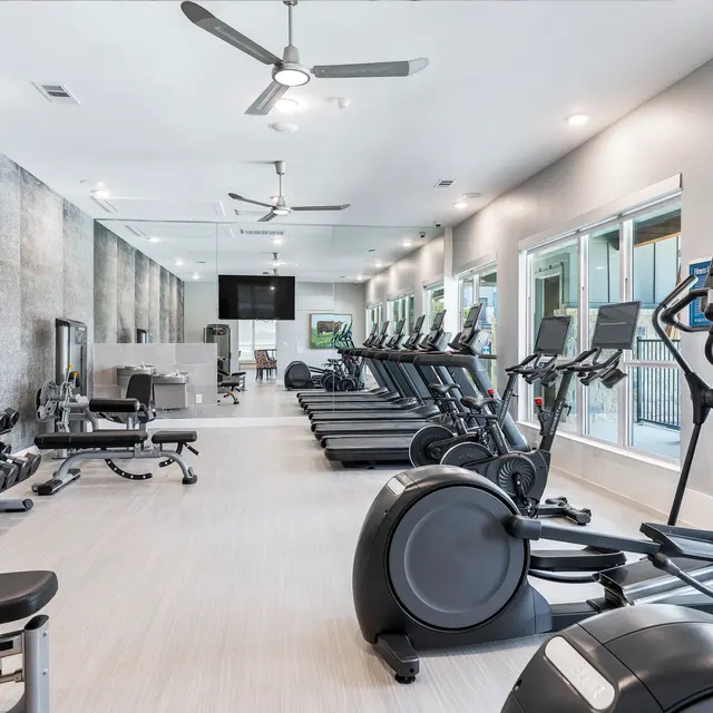 A modern gym interior featuring various exercise equipment including treadmills, ellipticals, and free weights. The space is well-lit with large windows and has a clean, contemporary design.