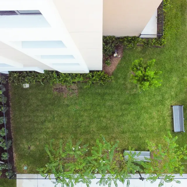 Aerial view of a small green space surrounded by a building and paved areas. The lawn is well-maintained with patches of foliage and benches visible.