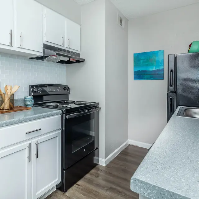 A modern kitchen featuring white cabinetry, a black stove and refrigerator, and a light gray countertop. A blue painting is hung on the wall, with utensils placed on a wooden cutting board.