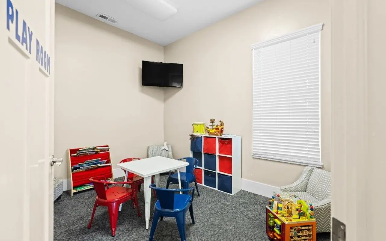 Children's Play Room A colorful playroom featuring a small table with chairs surrounded by various toys. There is a television mounted on the wall and a window with white blinds.
