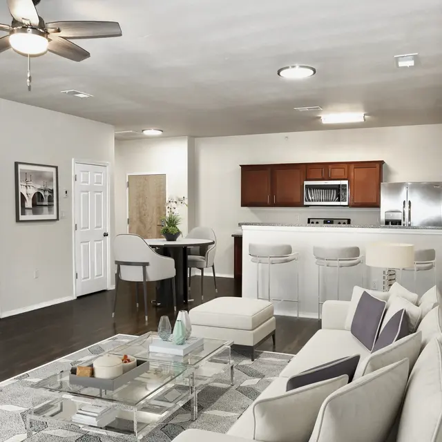 A modern living room featuring a white sofa, a glass coffee table, and an island bar with stools. The kitchen is in the background with dark wood cabinets and stainless steel appliances. The room has a ceiling fan and several doors leading to other areas.