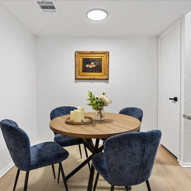 A cozy dining area featuring a round wooden table surrounded by four blue upholstered chairs. On the table, there are two candles and a vase with flowers. A piece of art hangs on the wall above the table. The walls are white and the floor is wooden.
