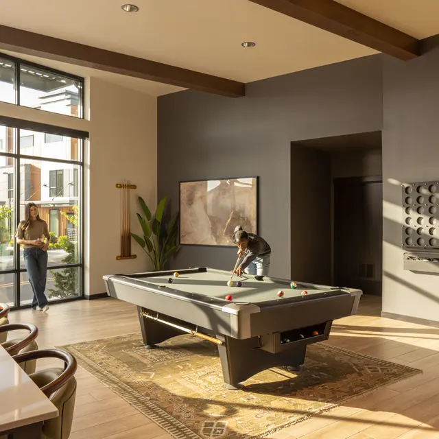 A spacious modern game room featuring a pool table, large windows allowing natural light, and a bar area with seating. One person is playing pool while another stands nearby, looking out the window. Decorative plants and artwork enhance the ambiance.