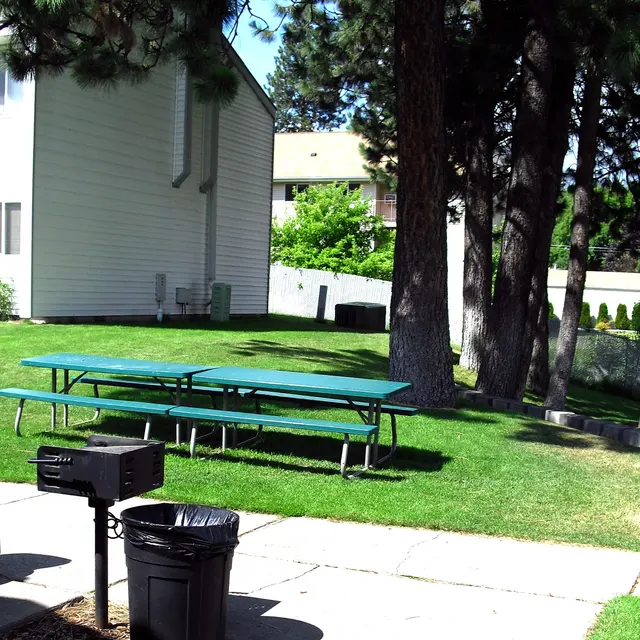 Outdoor Picnic Area A green picnic area featuring a few tables, a charcoal grill, and a trash can, surrounded by trees and a building in the background.