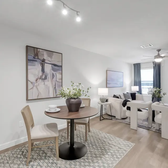 A modern dining area with a round wooden table and two chairs. In the background, a living area with a white sofa, decorative plants, and artwork on the walls. Bright and inviting atmosphere with natural light.