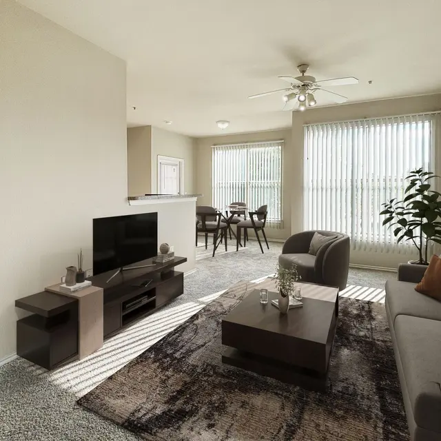A modern living room with a gray sofa, coffee table, and a television on a stand. Large windows with blinds provide natural light. There is a plant in the corner and a dining area visible in the background.