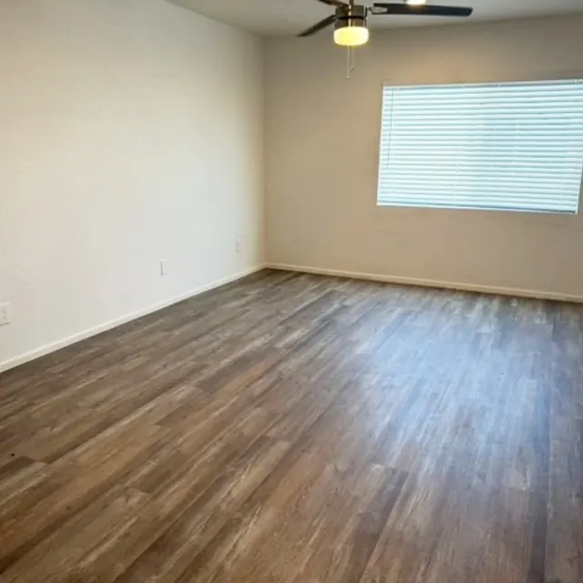 An empty living room with light-colored walls and wooden flooring, featuring a ceiling fan and a window with blinds.