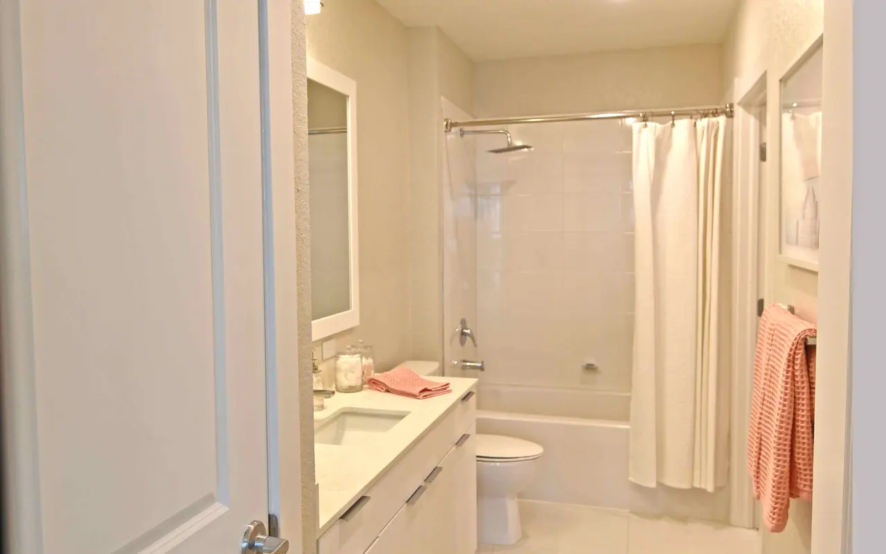 A modern bathroom featuring a spacious layout with white walls, a mirror above a sink, and a shower area with a curtain. A bath mat is on the floor, accompanied by a towel rack with a pink towel.