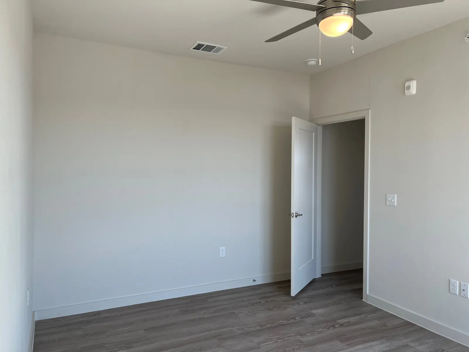 Empty Room A well-lit empty room with light-colored walls and wooden flooring. A door is slightly opened on the right side, and there is a ceiling fan above.