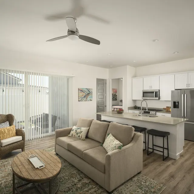 A modern living room with a beige sofa, a wooden coffee table, and a comfortable armchair. There is a fan on the ceiling and a kitchen area in the background with white cabinets and stainless steel appliances. Large sliding glass doors let in natural light, revealing a patio outside.