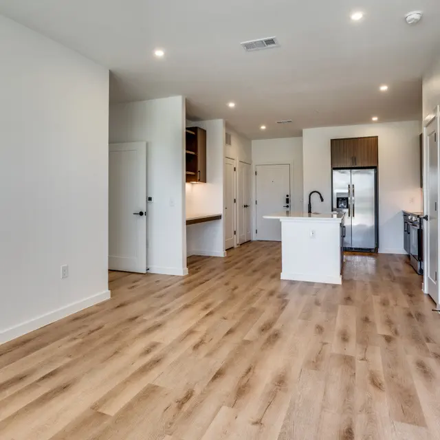 Spacious modern apartment interior with wooden flooring and an open layout, featuring a kitchen area and doors leading to other rooms.