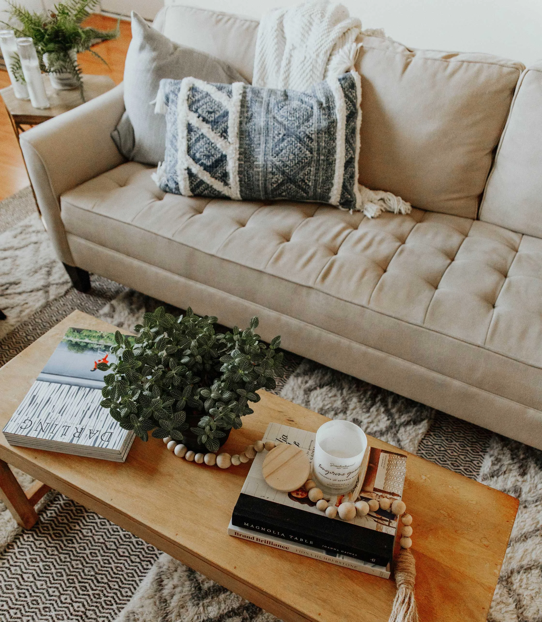 Cozy Living Room Setup A cozy living room scene featuring a beige sofa adorned with blue patterned pillows. A wooden coffee table is topped with books, a small potted plant, a candle, and decorative beads. The area is complemented by a textured rug and some greenery in the background.