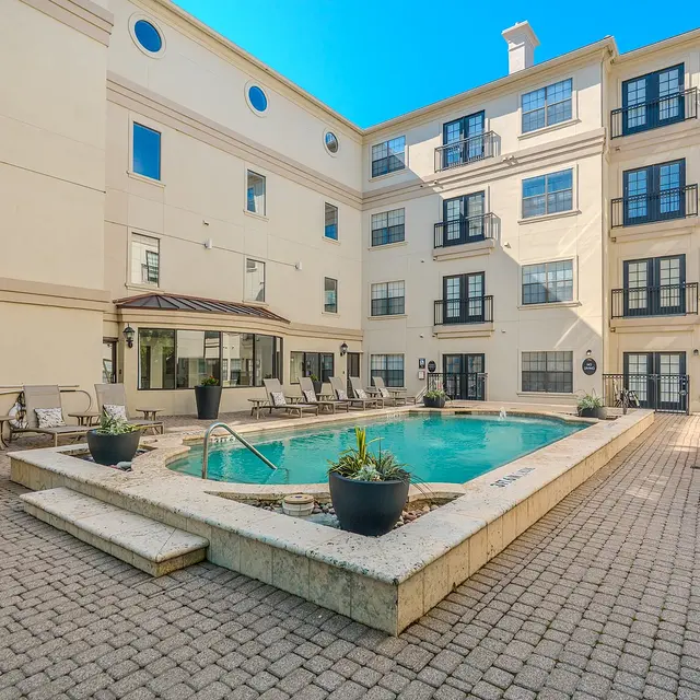 A well-maintained outdoor pool area surrounded by a paved courtyard, with loungers and a seating area, flanked by multi-story residential buildings.