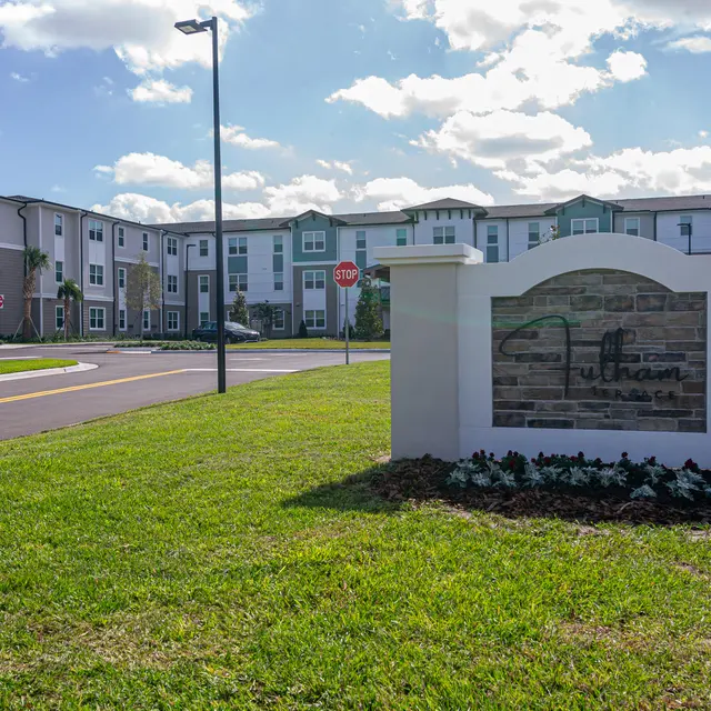 A large sign for Fulton apartment community in a green grassy area, with modern apartment buildings in the background under a partly cloudy sky.
