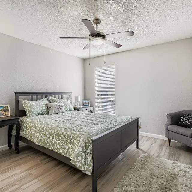 Cozy Bedroom Interior A cozy bedroom featuring a bed with a green patterned comforter, a gray accent chair, a small table, and a ceiling fan. There are images on the table and a window with blinds.