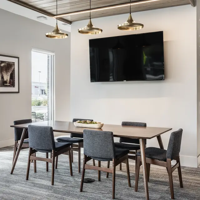 A modern meeting room with a large table and gray upholstered chairs, illuminated by pendant lights. There is a large screen on the wall and windows providing natural light.