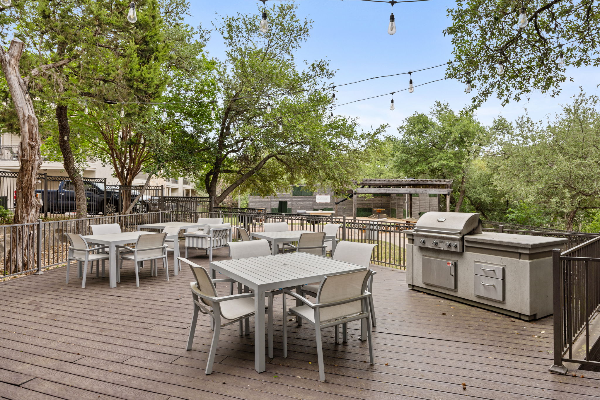 A spacious outdoor dining area with tables and chairs, set among trees and greenery, featuring a barbecue grill to the side.