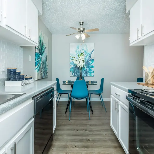 A modern kitchen with white cabinets, stainless steel appliances, and a dining area featuring blue chairs and a floral artwork on the wall.