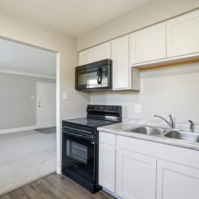 A modern kitchen with white cabinets, a black microwave and stove, and stainless steel sinks. The kitchen opens into a carpeted living area with light gray walls.