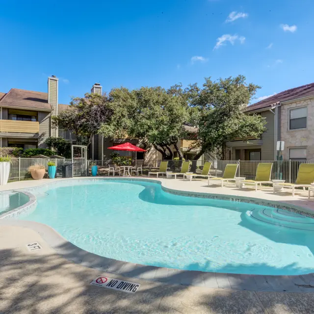 Outdoor swimming pool surrounded by lounge chairs with shade umbrellas and residential buildings in the background.