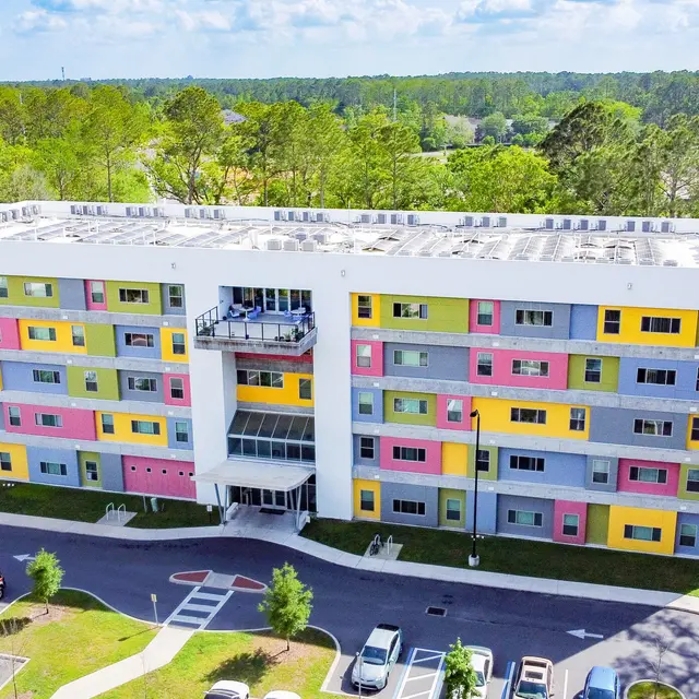A modern, multi-storied building with a vibrant facade featuring various colors like pink, yellow, and blue, surrounded by green trees and a parking lot.