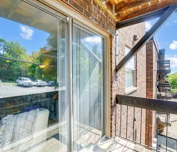 A balcony with sliding glass doors, featuring a view of trees and parked cars below. The floor is wooden and the ceiling is partially covered with beams.