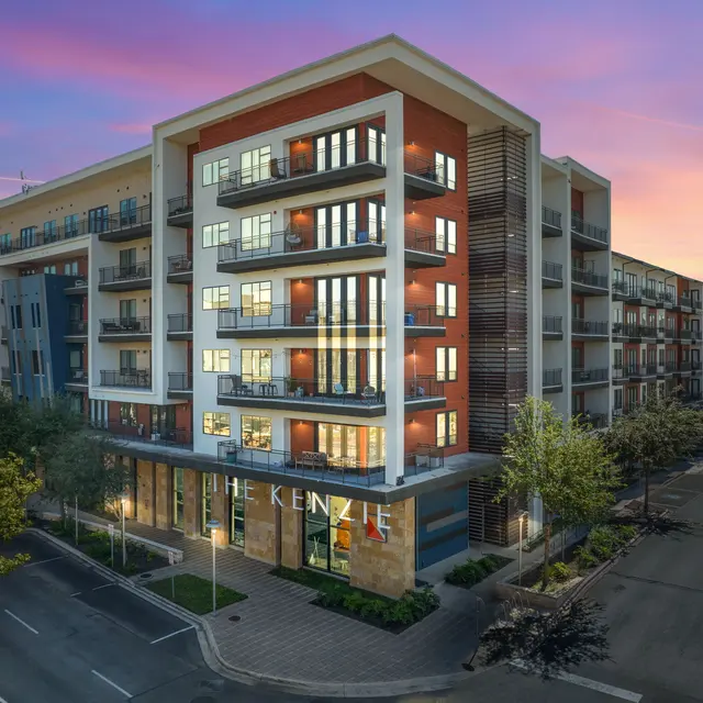 Modern apartment building with multiple balconies and large windows, surrounded by trees, at sunset.