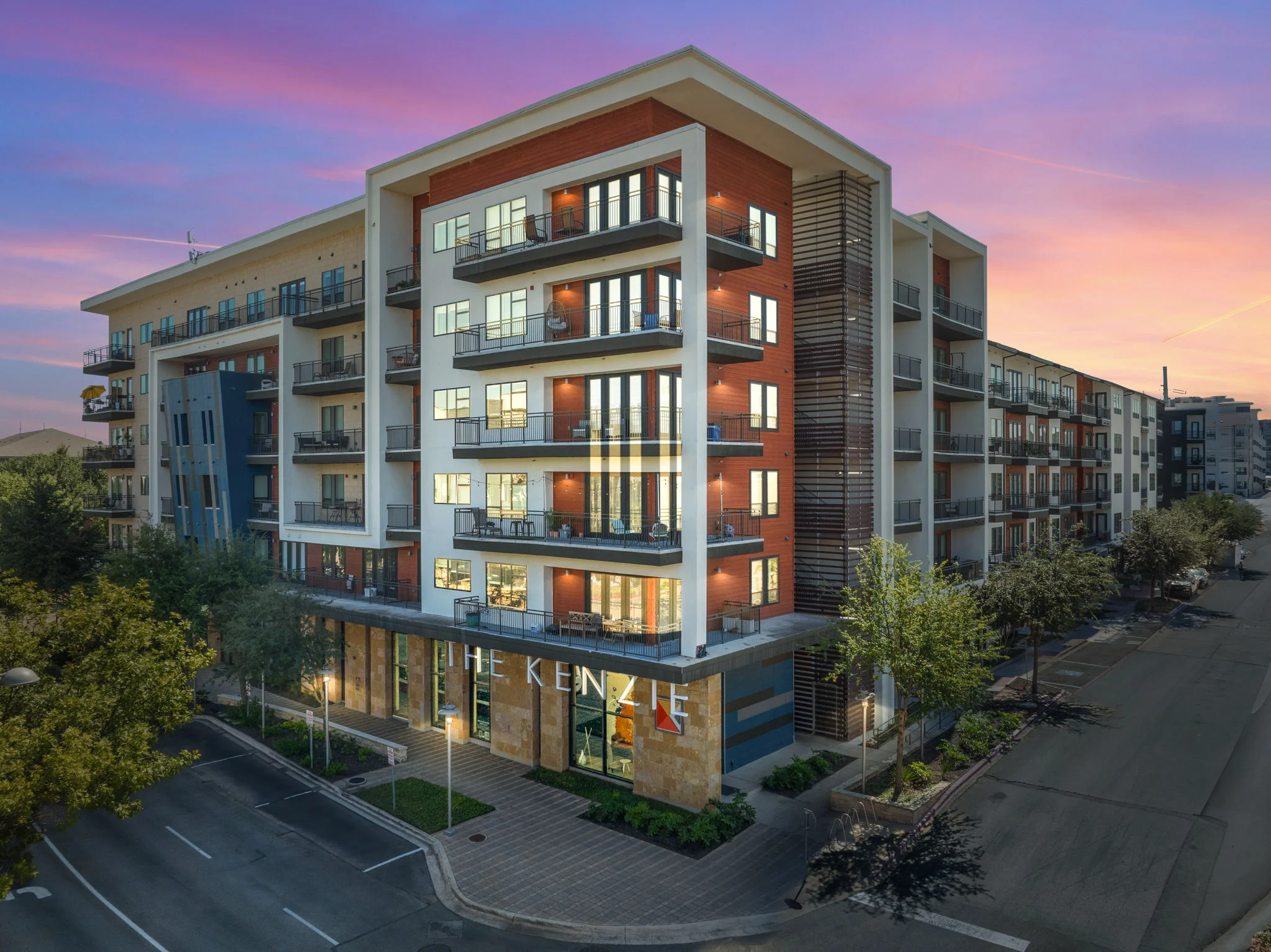 Modern apartment building with multiple balconies and large windows, surrounded by trees, at sunset.