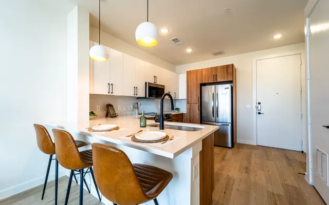A modern kitchen featuring a large island with bar stools, white cabinetry, and stainless steel appliances. The space is bright with natural light and has pendant lighting. There is a small green plant on the counter.
