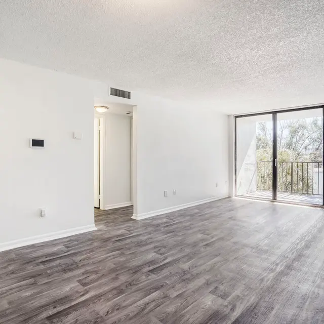 Modern Living Room Interior A spacious, empty living room featuring large windows that provide natural light, light-colored walls, and wood-style flooring. A doorway leads to a kitchen area visible in the background.
