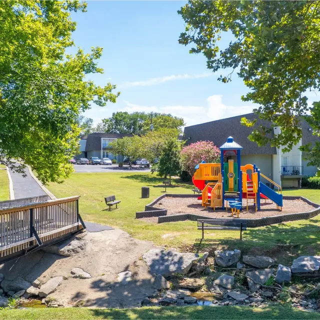Colorful Playground in Park A colorful playground with slides and climbing structures surrounded by grass and trees in a bright park setting. A wooden bridge leads to the playground area.