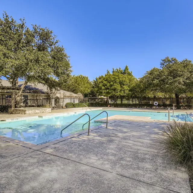 A serene swimming pool area surrounded by lush green trees and clear blue skies. The pool features clean water and a spacious patio with steps leading into the water.