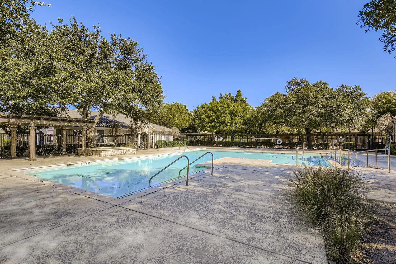 Serene Swimming Pool Area A serene swimming pool area surrounded by lush green trees and clear blue skies. The pool features clean water and a spacious patio with steps leading into the water.