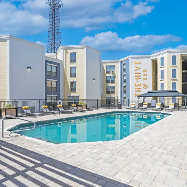 A spacious outdoor pool area surrounded by modern apartment buildings. Lounge chairs are placed around the pool, and there's a shaded seating area with a canopy. A tall communication tower is visible in the background against a bright blue sky with fluffy clouds.