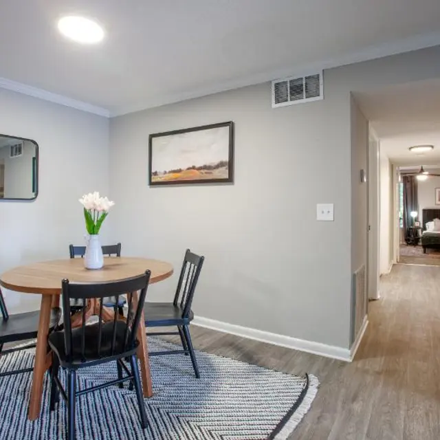 A cozy dining area featuring a round wooden table with four black chairs. A vase with white flowers sits on the table. The walls are painted light gray, and a large mirror hangs on the wall beside a framed landscape painting. A woven rug is under the table. In the background, a hallway leads to other rooms.