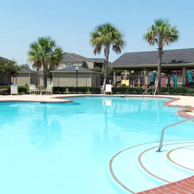 A clear blue swimming pool surrounded by lounge chairs and palm trees, with residential buildings in the background.
