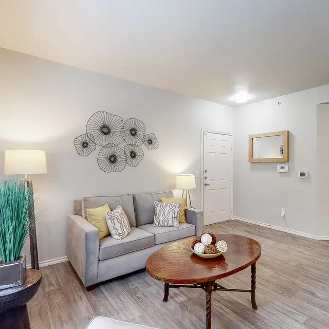 A modern living room featuring a gray sofa with decorative pillows, a wooden coffee table with decorative items, a tall green plant, and accent lighting on a side table. The walls are painted in light neutral tones and there is a decorative wall piece.