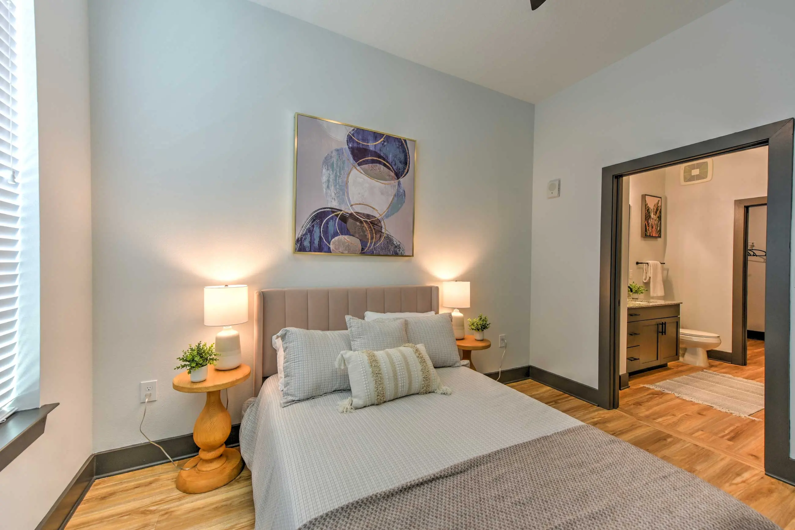 Modern Bedroom Interior A cozy bedroom featuring a light gray bed with decorative pillows, two round wooden side tables with lamps, and a decorative wall art piece. The room has hardwood flooring and a doorway leading to a bathroom.