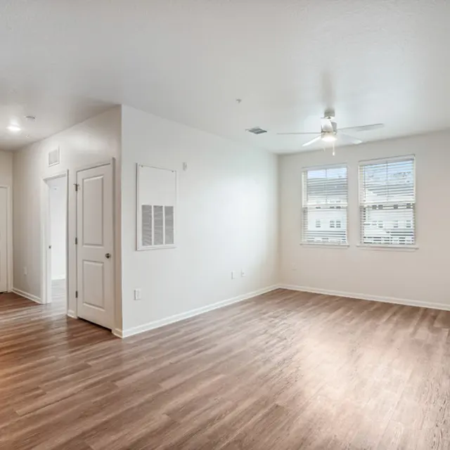 Bright, empty living room with hardwood floors, large windows, and a ceiling fan, adjacent to a kitchen area.