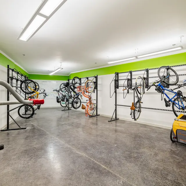 A bike storage area featuring multiple bicycles hung on wall racks, a bike repair station in the foreground, and a clean, well-lit environment with green accents.