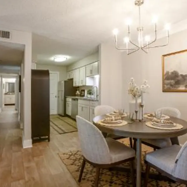 A dining area featuring a round table set for four with a chandelier overhead and an adjacent kitchen area. The room is warmly lit with light-colored walls and wooden flooring, creating a cozy atmosphere. A hallway is visible in the background, leading to other rooms.